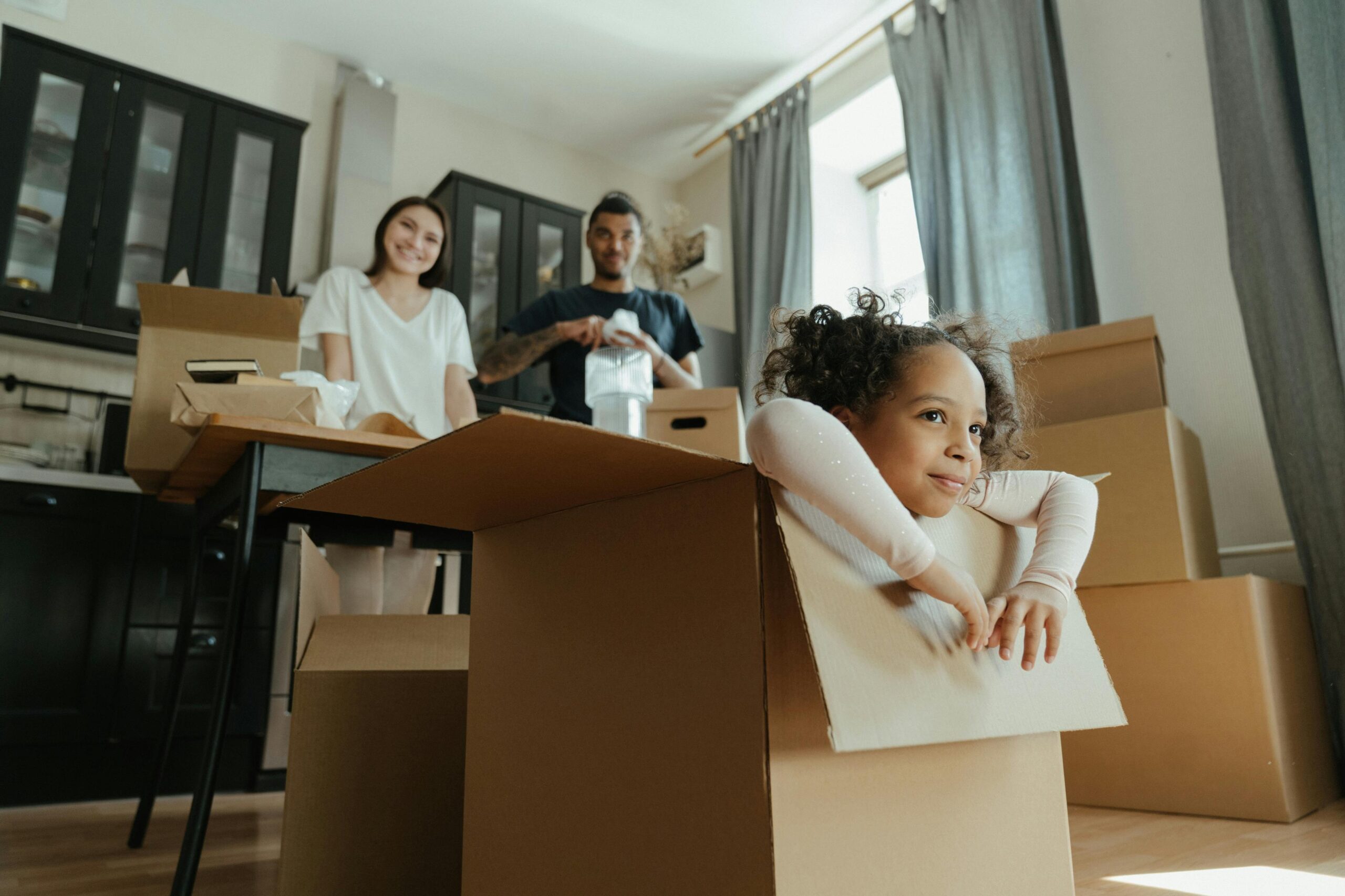 Family packing and preparing for a move in Platte Woods, MO, with a young child sitting in a cardboard box, smiling as they get ready for their new home. Perfect for those looking to sell their home fast to cash buyers in Platte Woods, MO.